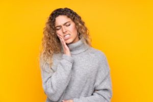 Woman in gray sweater against a yellow background holding the side of her mouth in pain