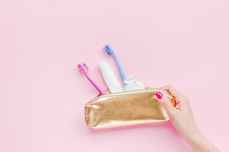 Woman's manicured hand holding small gold pouch with two toothbrushes, toothpaste, and floss spilling out over baby pink background